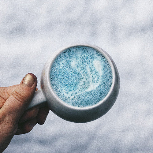 a hand with gold nail polish is holding a mug over top snow. Inside the mug is a bright blue and bubbly liquid that is laced with white from milk.