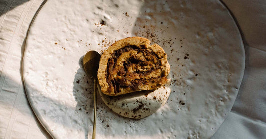 a large white plate sits on top of bedding with a round tan coloured cinnamon roll in the middle and a small golden spoon beside it ready to eat