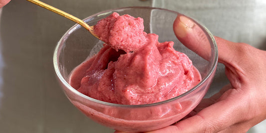 a close up of a clear bowl filled with pink sorbet in someones hands who is lifting a gold spoon filled with sorbet