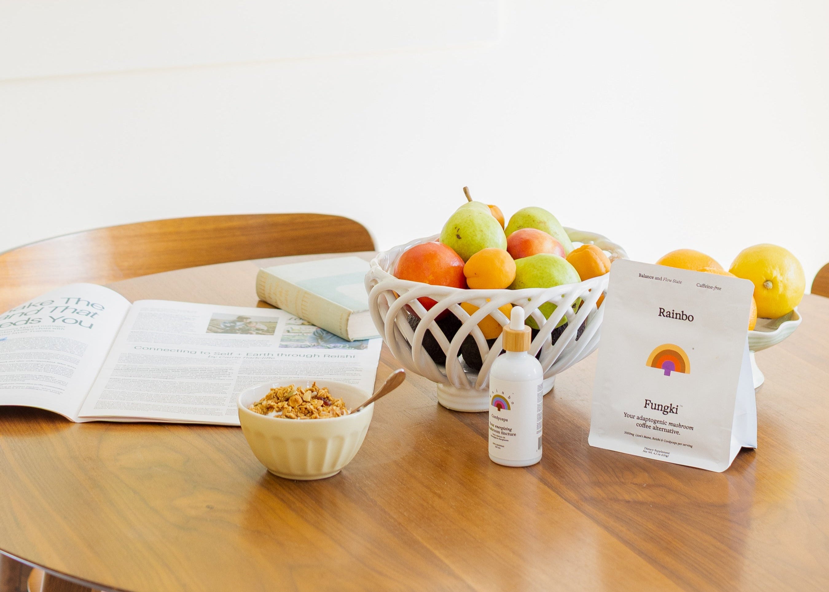 Table with a bowl of fruit, a Rainbo Cordyceps tincture bottle, a bowl of yogurt, a bag of Fungki mushroom coffee alternative and an open newspaper on a white background.