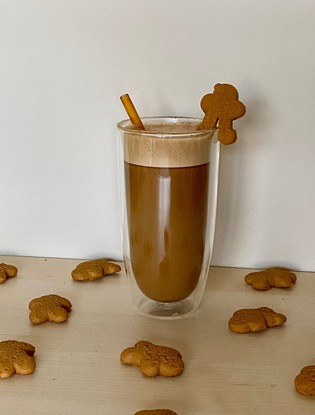 a tall glass tumblr mug is filled with a brown coffee with frothed milk at the top. It is garnished with a gingerbread man on the rim of the glass 