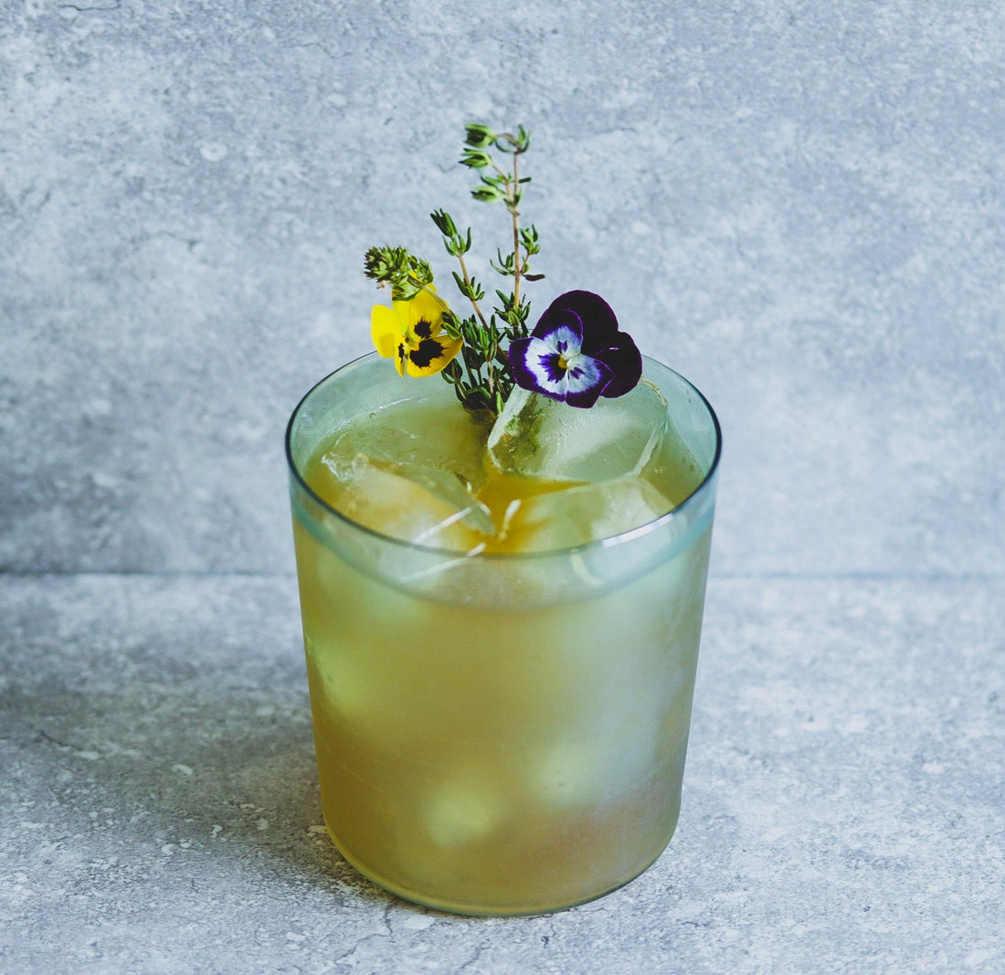 In the centre of a grey marbled background there is a frosted green glass filled with a yellow liquid and ice. It is garnished with two pansy flowers and a sprig of thyme