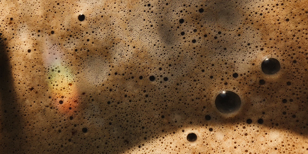 a very close up image of the bubbles from a frothed coffee. The coffee varies in shades of brown and you can see bubbles in the midst of popping. There is also a small rainbo in the bottom left corner of the image as a result of the sun