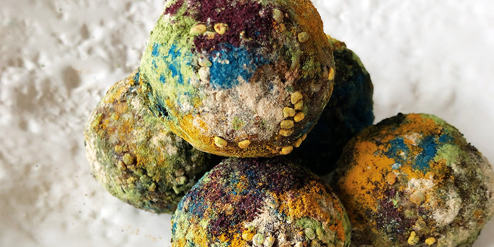 On a textured white plate, there is a close up of five balls stacked onto one another. Each ball has been rolled in various powders such as spirulina, blue majik, tumeric, cinnamon, and bee pollen. Each ball features the colours blue, green, orange