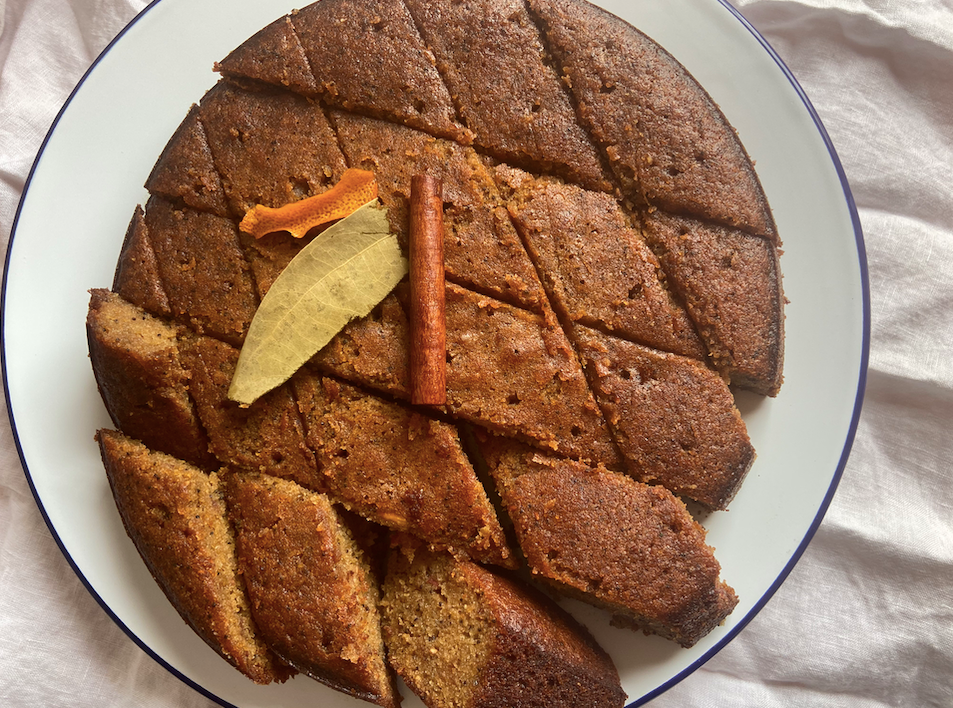 round brown cake on a plate cut into diamond shapes with a cinnamon stick on top and a blood orange peel