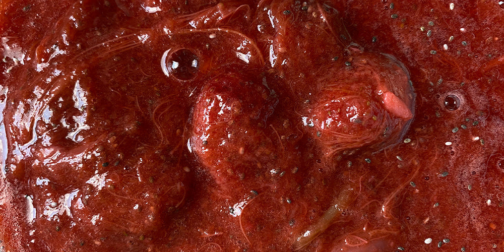 close up strawberry rhubarb jam with a deep red colour. The jam has some bubbles in it from the stove. Jam has some chia seeds in it and looks delicious.
