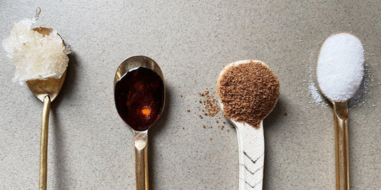 4 spoons are lined up on a sandy grey background the first one has honey in it, the second has maple syrup, the third coconut sugar and the fourth is refined white sugar
