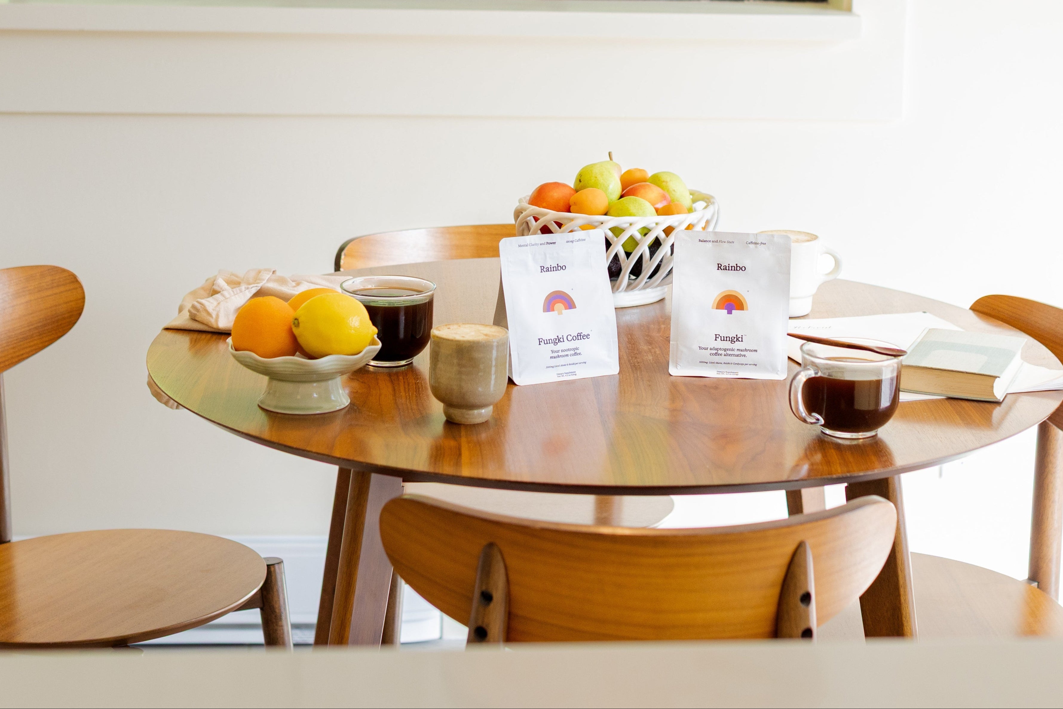 Round wooden table with chairs, fruit bowl, coffee cups, Fungki and Fungki Coffee gusset bags and books in a bright room.